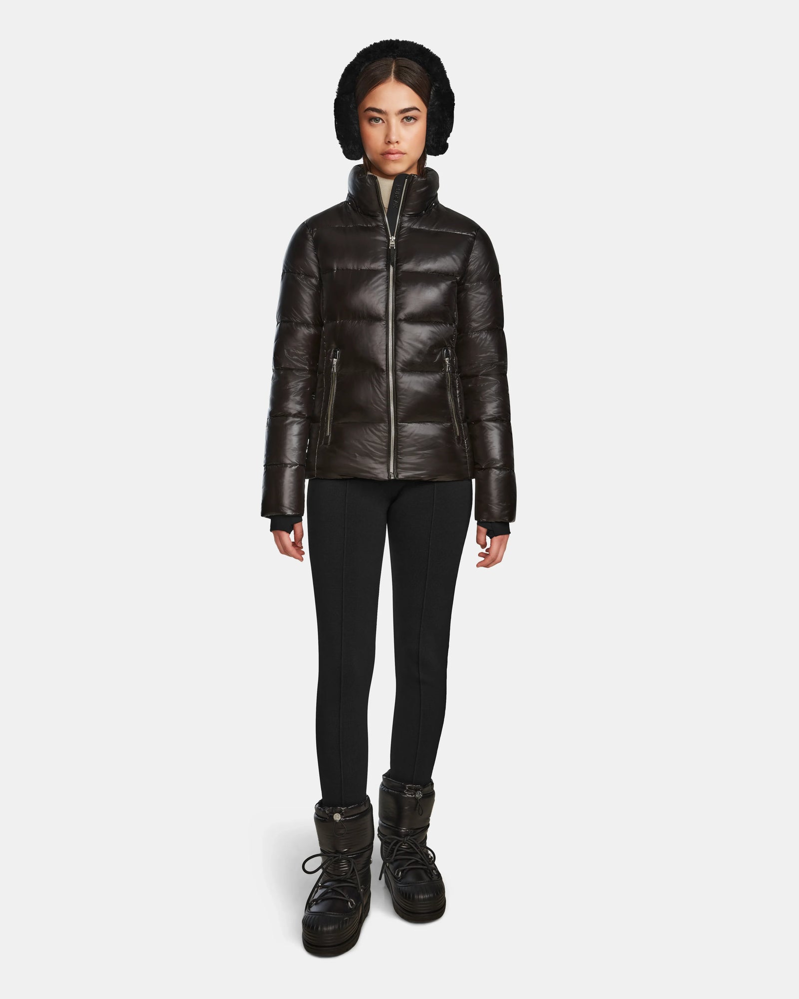 A woman stands facing forward in a shiny black Rudsak MIRRY S - L123545 womens quilted down coat, paired with black leggings, winter boots, and a fur-lined hat with ear flaps against a plain white background.