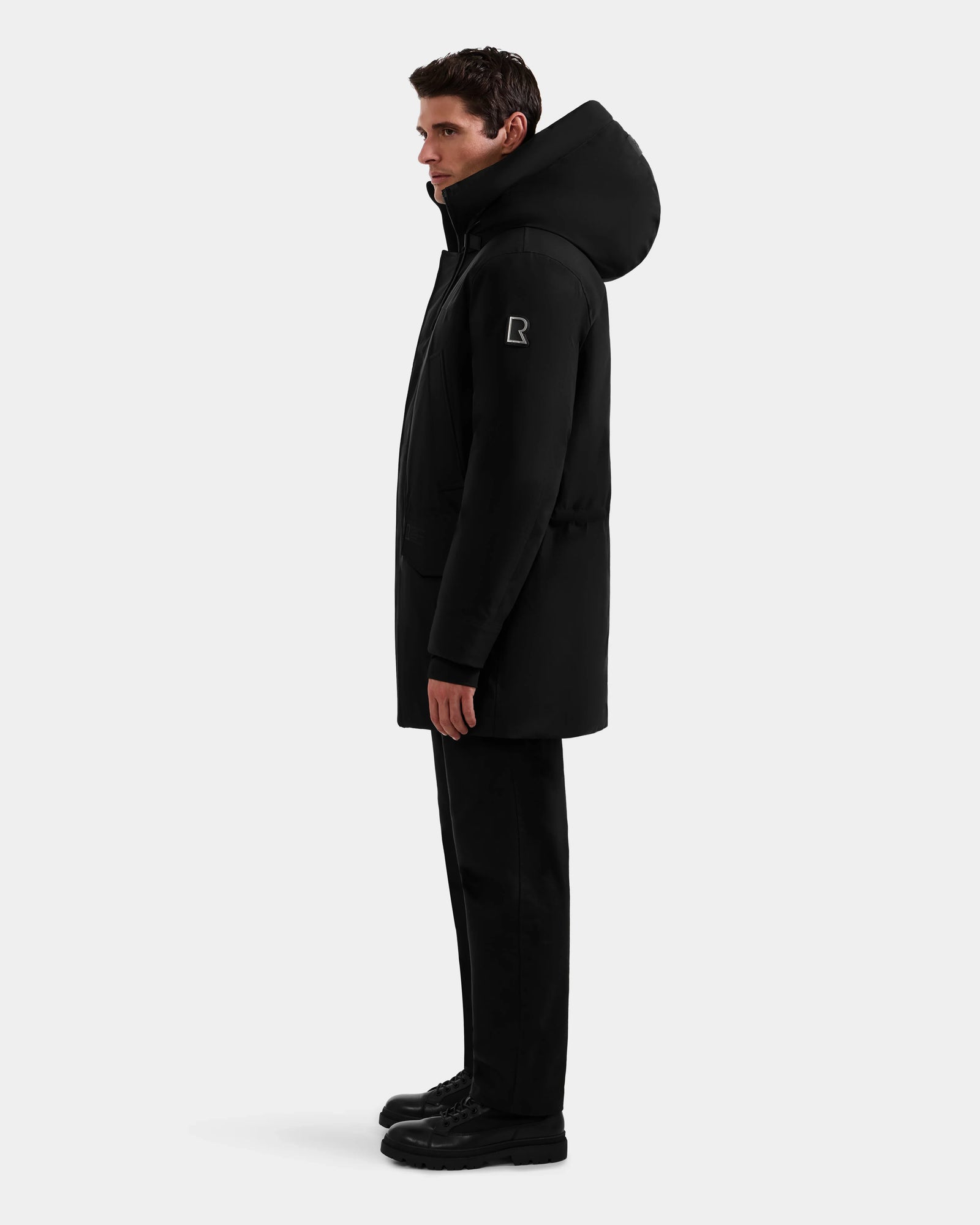 A person stands in profile wearing the Rudsak MITCHELL - M125513 Black men’s down parka with a sleeve patch, paired with black pants and shoes, against a light gray background. The parka is filled with RDS-certified down for warmth.