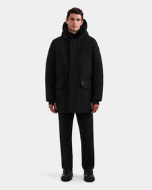 A man faces forward against a plain white background, wearing the Rudsak MITCHELL - M125513 Black men’s down parka with RDS-certified down, paired with black pants and black shoes.