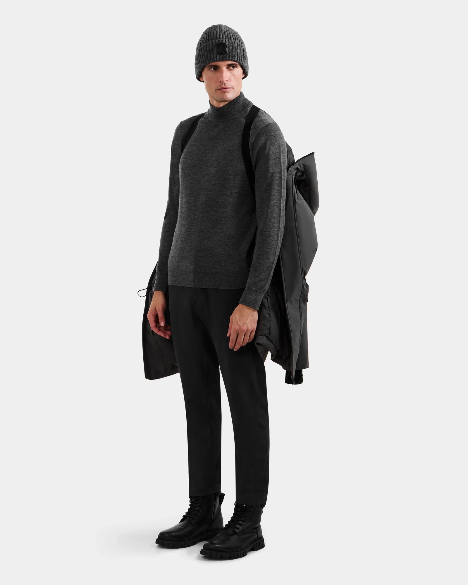 A person stands against a plain background wearing the Rudsak MITCHELL - M125513 Grey knit beanie, paired with a gray sweater, black pants, black boots, holding a men’s down parka over one shoulder and a black bag on their back.