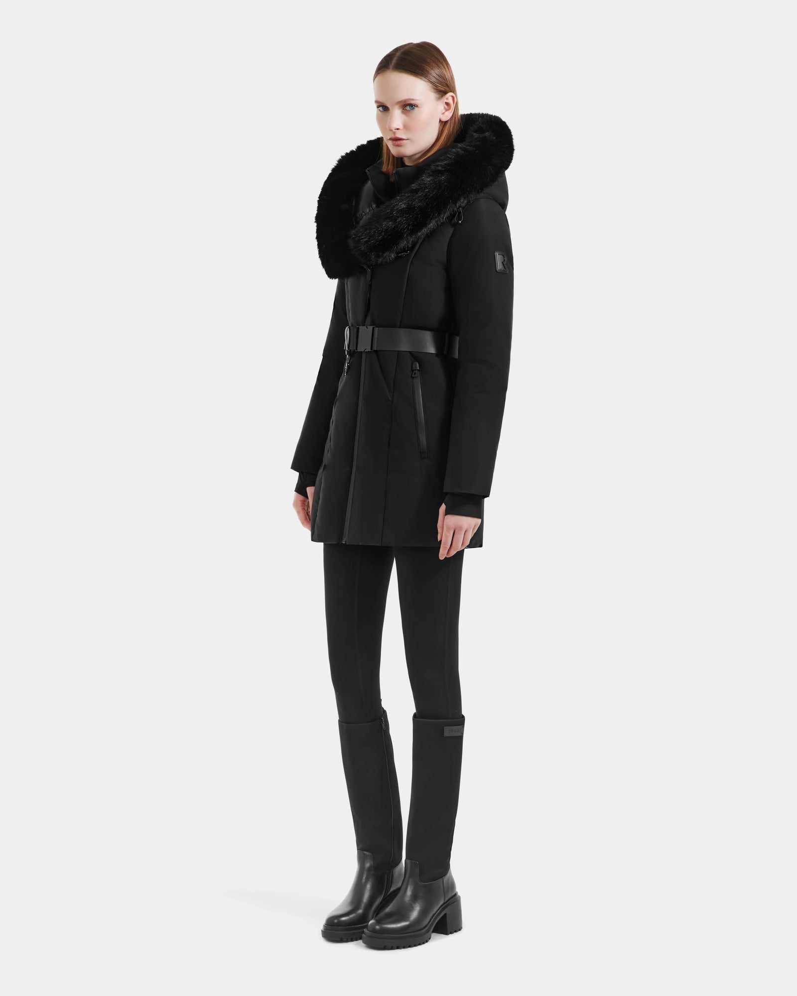 A woman models the Rudsak MODA M - L125555 Black coat, featuring a large fur collar and vegan leather belt, with black leggings and knee-high boots. She stands against a white background with straight, shoulder-length hair.
