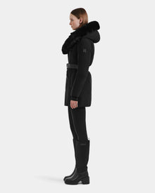 A woman stands in profile against a light background, wearing the Rudsak MODA M - L125555 Black winter coat with a large faux fur-trimmed hood, black leggings, knee-high boots, and a vegan leather belt at the waist.