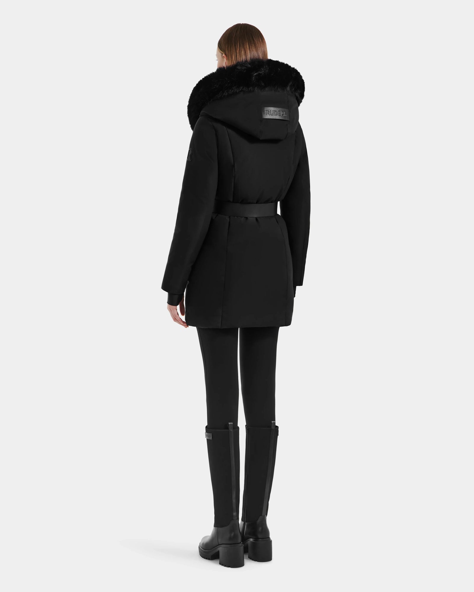 A woman faces away, wearing the Rudsak MODA M - L125555 Black winter coat with a large furry hood, vegan leather belt, black leggings, and knee-high black boots against a plain light background.