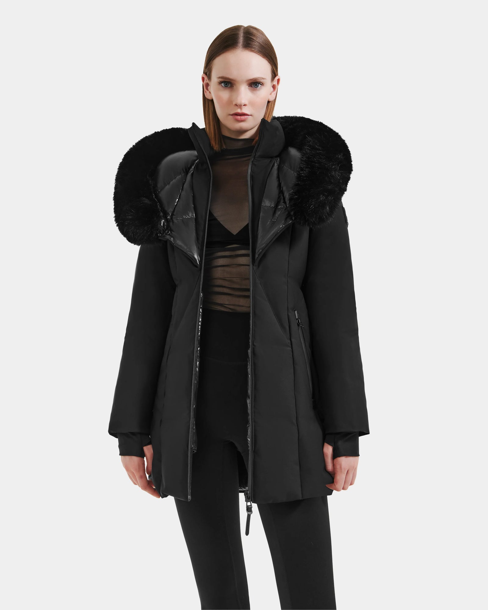 A woman faces forward against a plain white background wearing the Rudsak MODA M - L125555 Black winter coat with a large, fluffy fur-lined hood, paired with a sheer black top and black leggings.