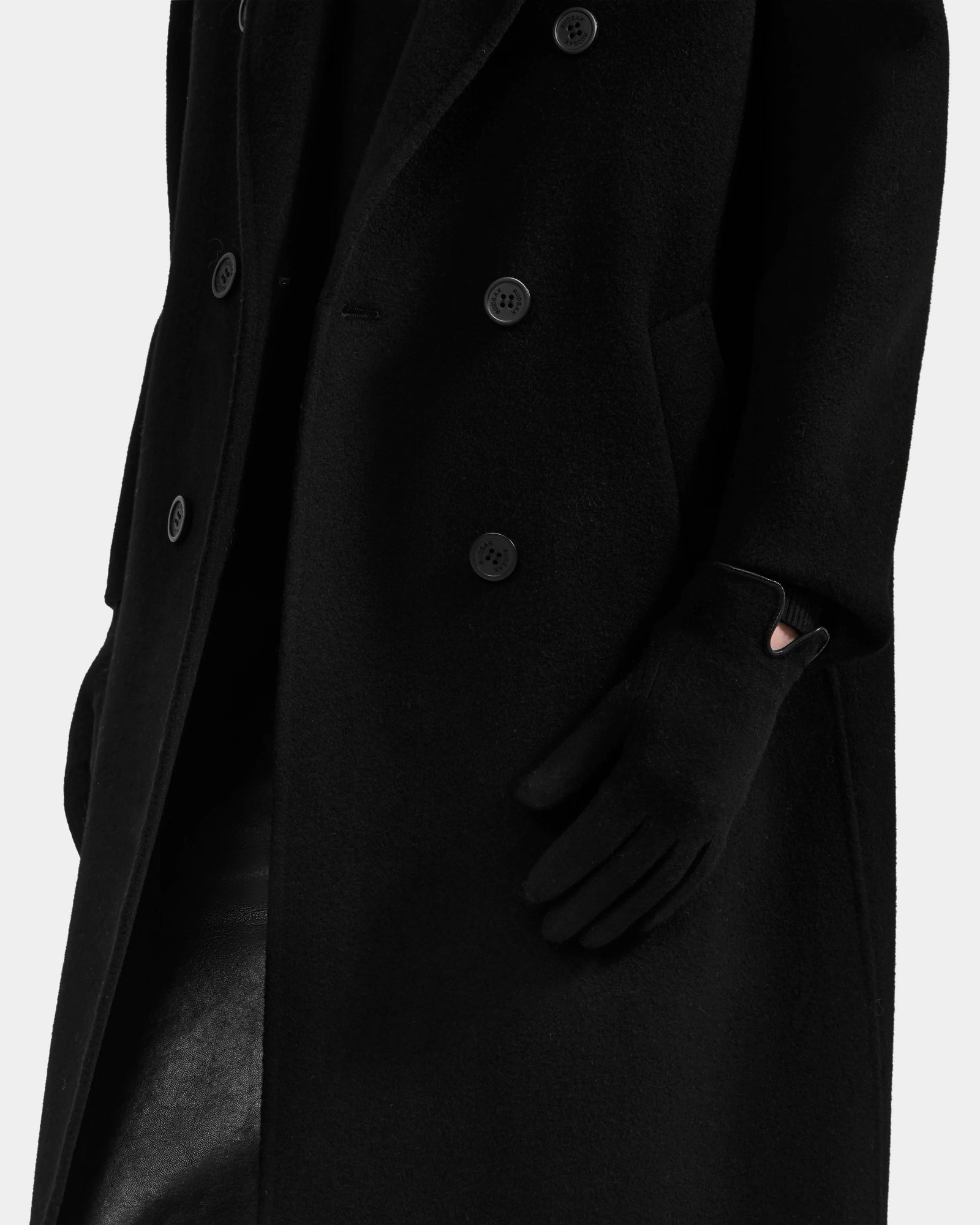 A person wears the Rudsak OLGA - L425541 Black long, double-breasted coat with women's wool gloves, one hand tucked in a pocket. The plain white background highlights this cold-weather essential look.
