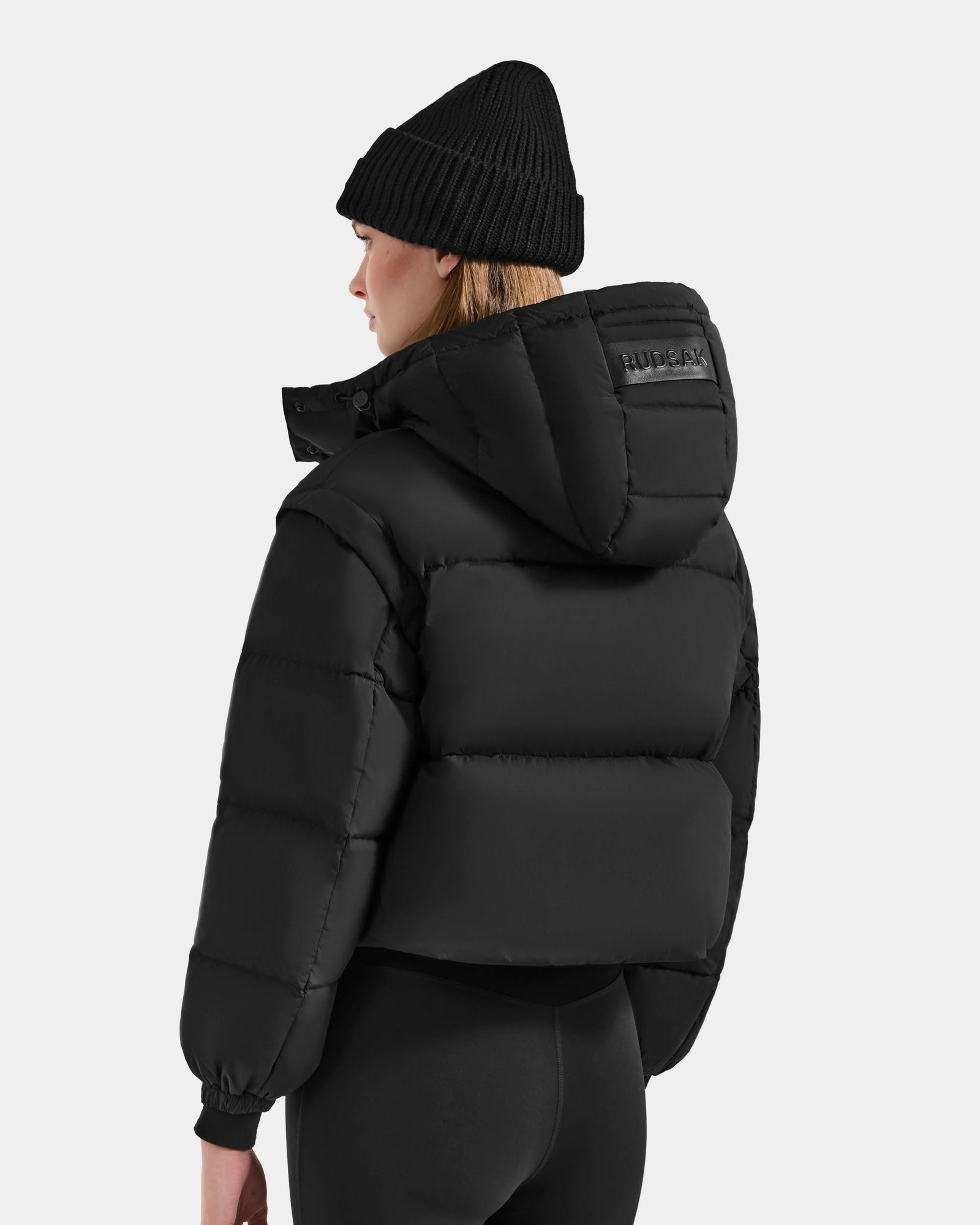 A woman with straight blonde hair wears the Rudsak OPRAH - L125546 Black cropped down puffer jacket with a hood, paired with a black beanie and leggings. She stands facing away, showcasing the back of the outfit against a plain light background.