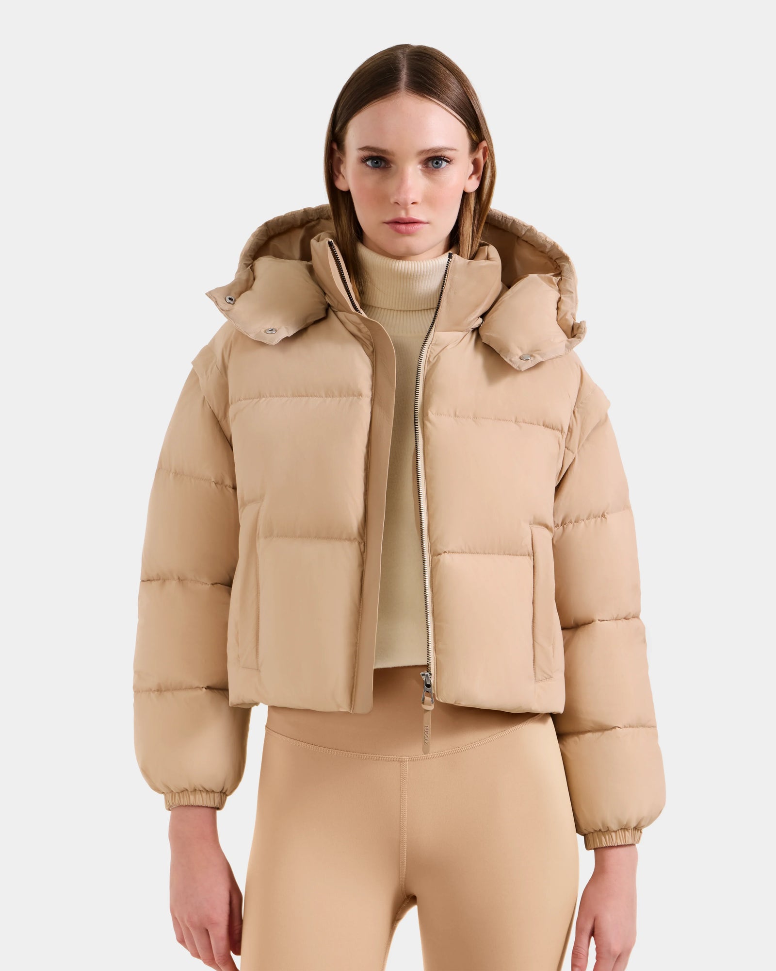 A woman stands facing forward against a plain white background wearing the Rudsak OPRAH - L125546 Vanilla women's cropped down puffer with removable sleeves, paired with a beige turtleneck and matching leggings.