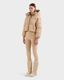 A woman faces forward wearing the Rudsak OPRAH - L125546 Vanilla cropped down puffer jacket with removable sleeves, paired with matching beige leggings and knee-high winter boots, set against a plain white background.