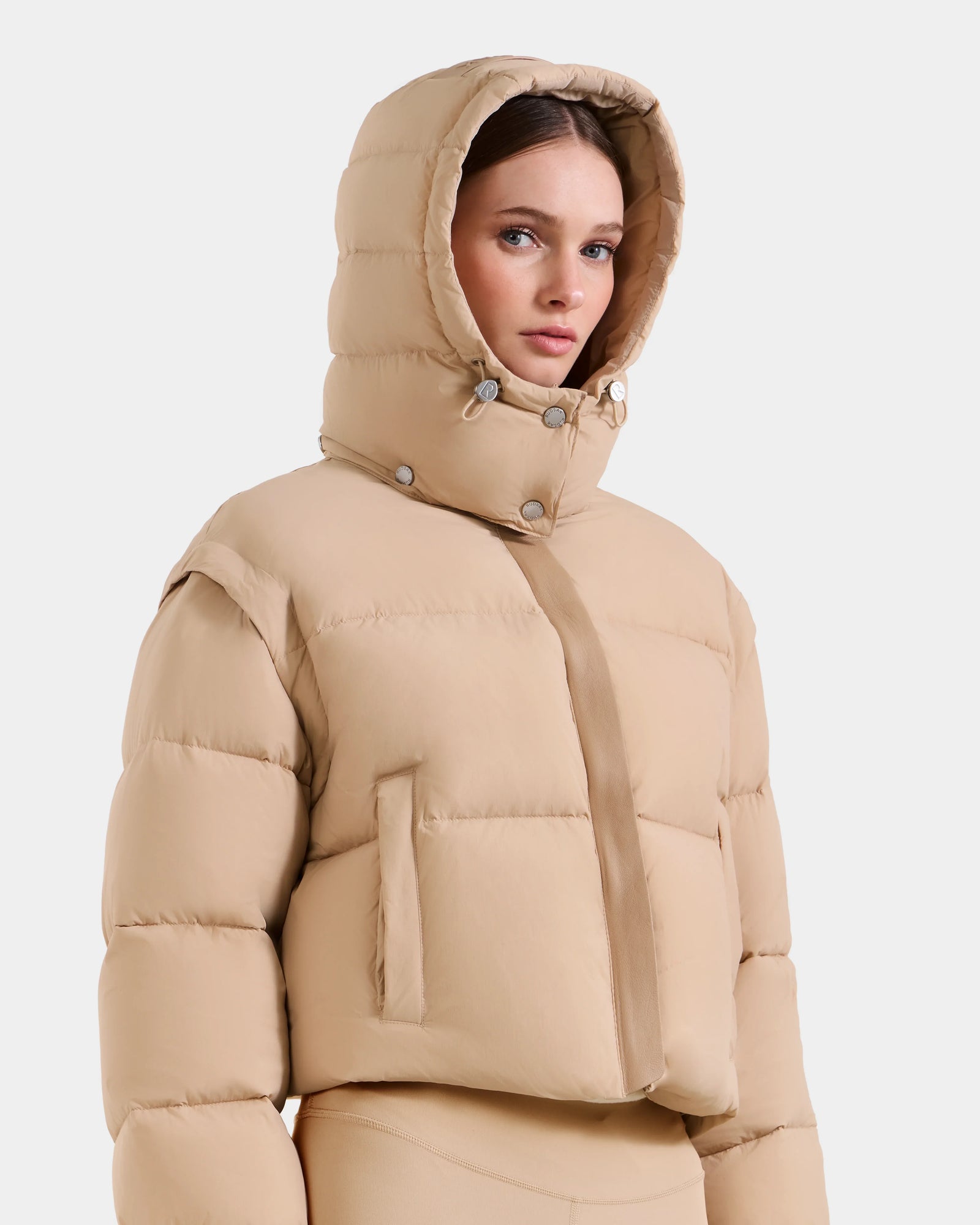 A woman in the Rudsak OPRAH - L125546 Vanilla beige cropped down puffer with hood, large horizontal quilting, and snap buttons near the collar. Made with RDS-certified down, this jacket stands out against a plain white background.