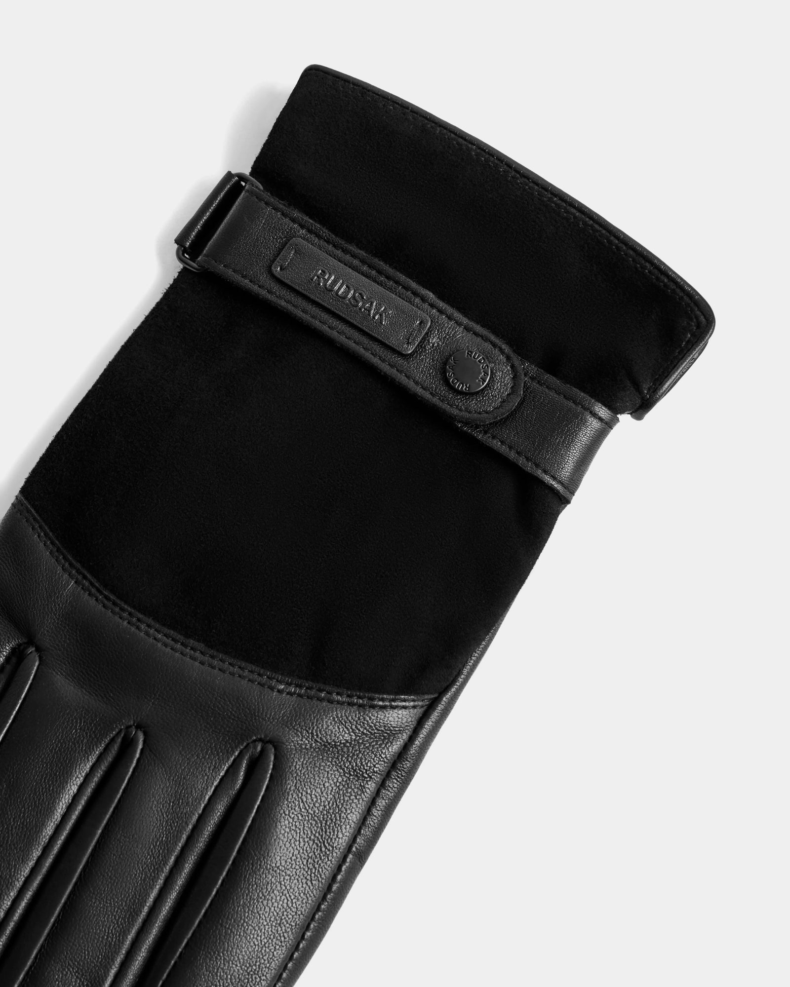 A close-up of the Rudsak PARKER - M425502 Black men’s leather glove, showing its strap with embossed text and a metal snap button, photographed on a light gray background.