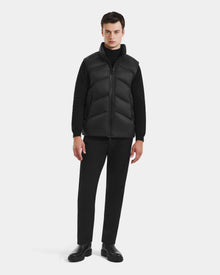 A man faces forward in a Rudsak RICHARDSON - M125527 Black men's hooded vest over a black turtleneck, pants, and shoes against a white background. The vest provides water-repellent protection and lightweight warmth for comfort.