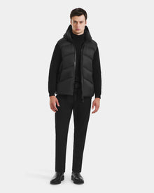 A man faces forward in the Rudsak RICHARDSON - M125527 Black men’s hooded vest over a black turtleneck, black pants, and shoes. Its lightweight warmth and water-repellent finish stand out against a plain light background.