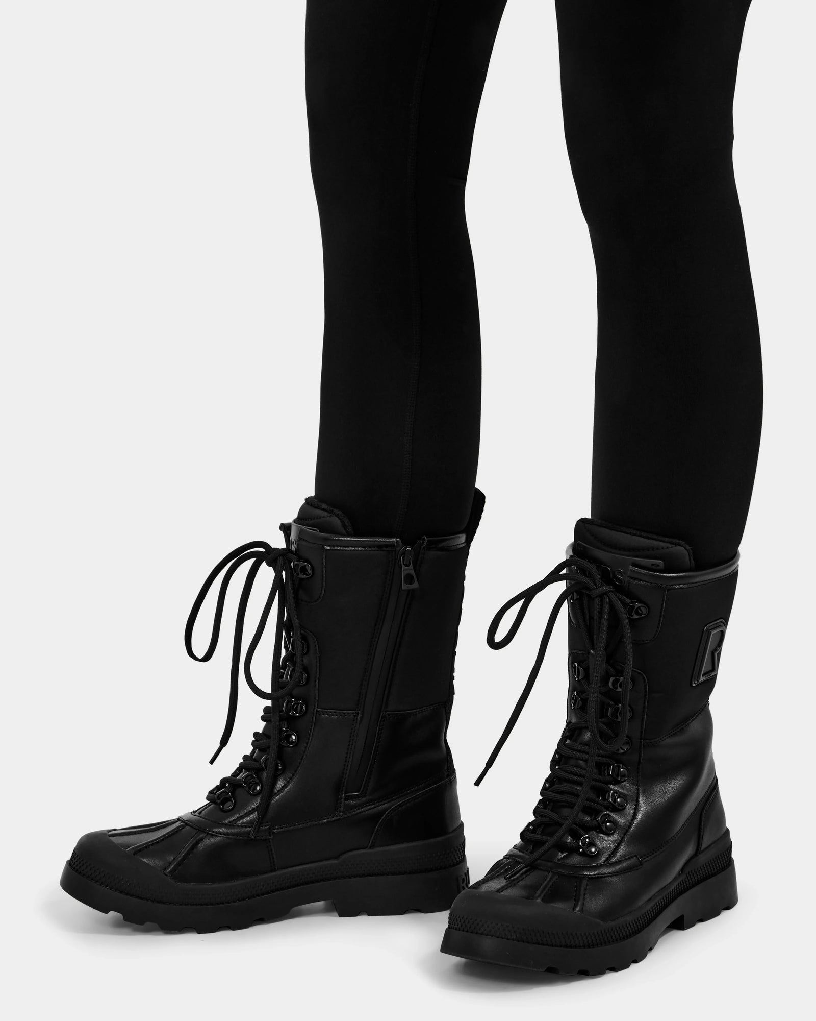 A person wearing Rudsak’s RITA - L222502 Black lace-up combat boots with side zippers and black leggings stands against a plain light background.