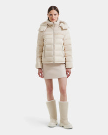 A woman faces forward against a white background wearing the Rudsak SANDRA - 8125612 Vanilla puffer jacket, a matching short skirt, and tall cream boots.