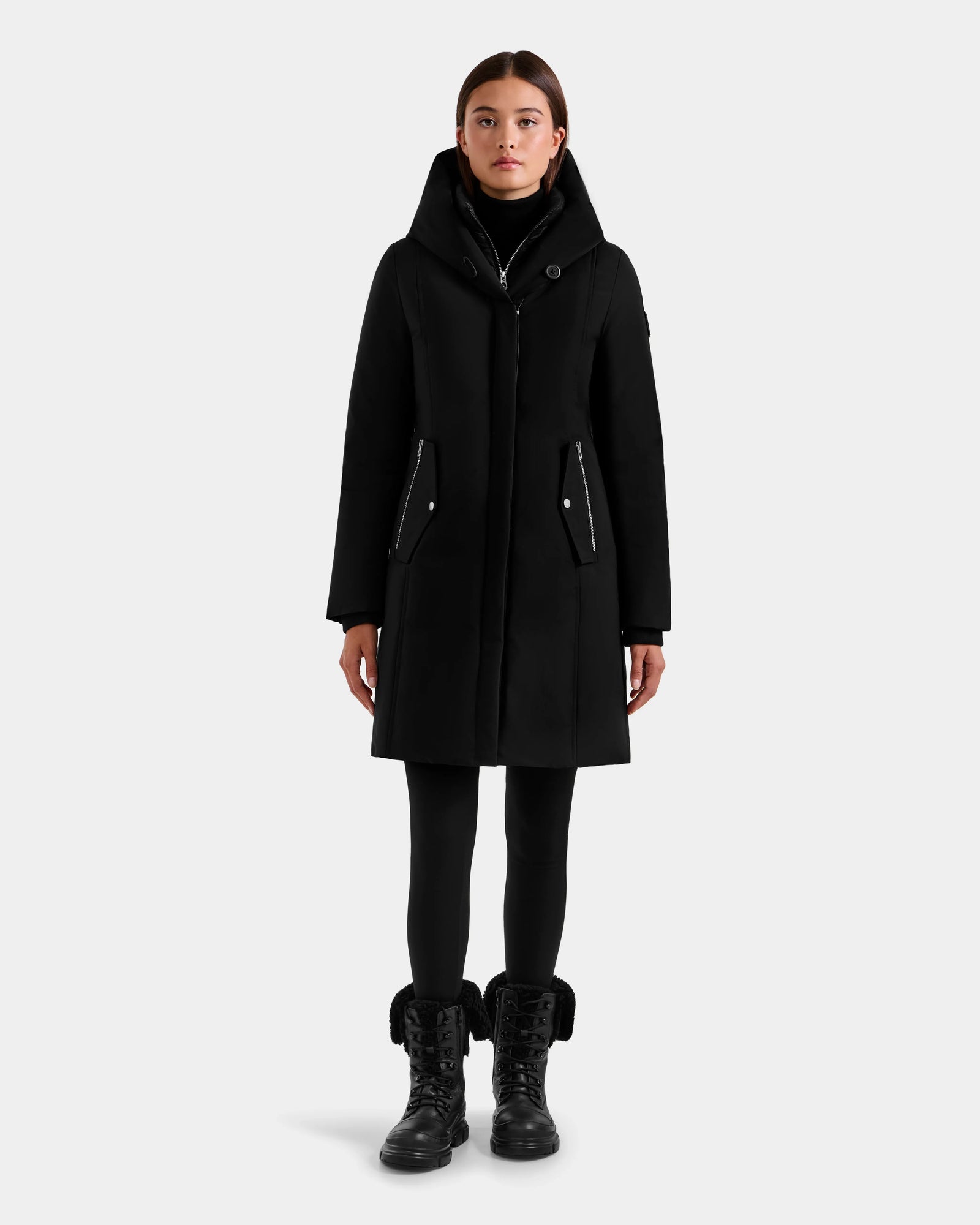 A person stands facing forward, wearing the Rudsak SARINA - L125550 Black women’s down parka with a high collar, black leggings, and black lace-up fur-trimmed boots against a plain light gray background.