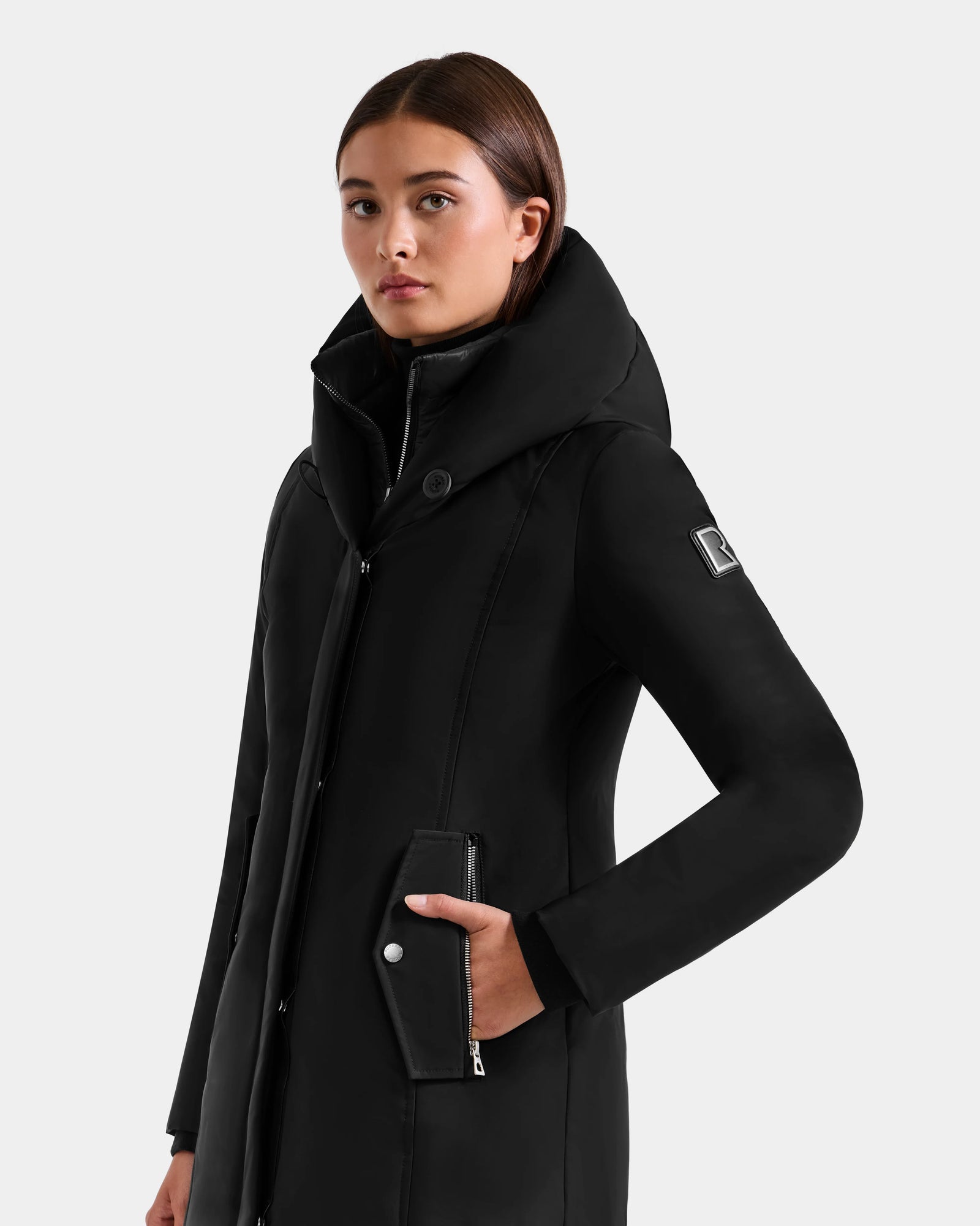 A woman wears the Rudsak SARINA - L125550 Black women’s down parka with a hood, high collar, and zippered front, standing against a light background and looking to the side with one hand in her pocket.