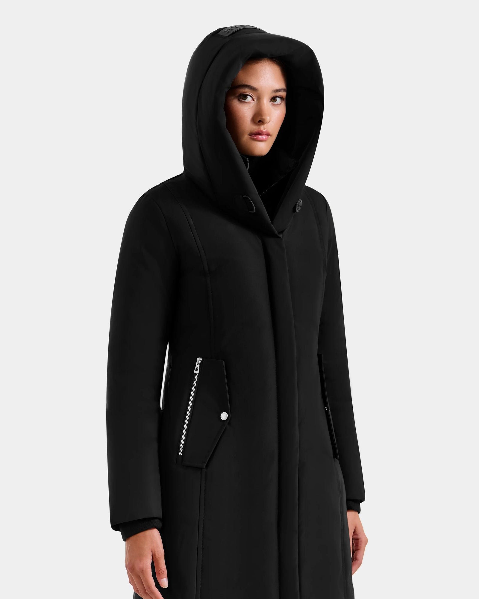 A person wears the SARINA - L125550 Black semi-fitted parka by Rudsak, featuring zippered pockets, standing against a plain light gray background with a neutral expression.