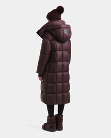 A person is seen from behind wearing the Rudsak SASHA - 8125686 Bordeaux women’s long down puffer coat with a hood, black leggings, a maroon pom-pom beanie, and matching boots.