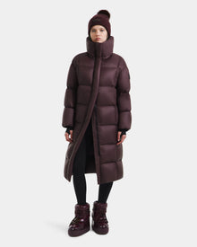 A person wearing the Rudsak SASHA - 8125686 Bordeaux long down puffer coat, matching boots, black leggings, and a maroon knit beanie stands against a plain white background.