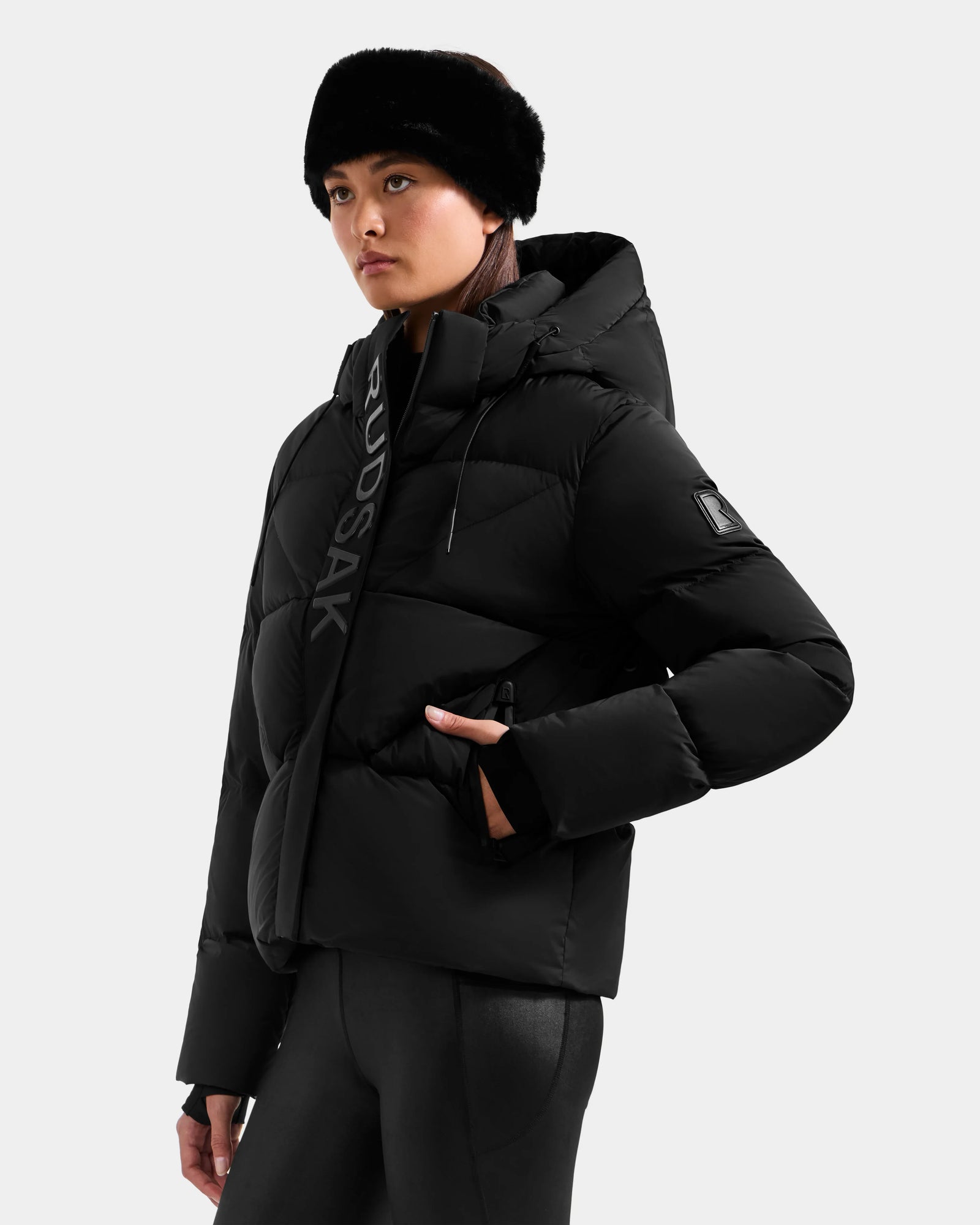 A woman wears the Rudsak STEFANIE M - L125526 Black puffer jacket, black leggings, and a black fur headband, standing against a light grey background with her hand in her pocket, looking to the side.