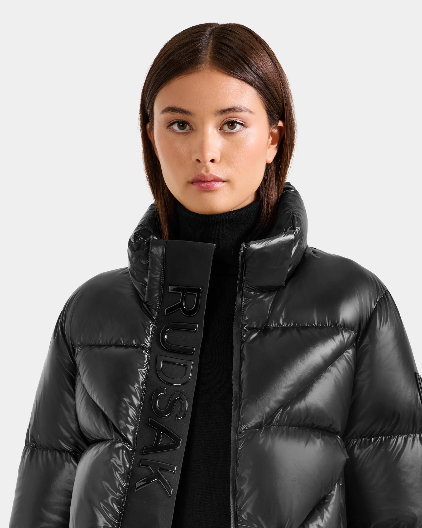A woman with straight brown hair models the Rudsak STEFANIE S - L125585 Black jacket, featuring a shiny, weather-resistant finish and "RUDSAK" on the collar, as she faces the camera against a plain light gray background.