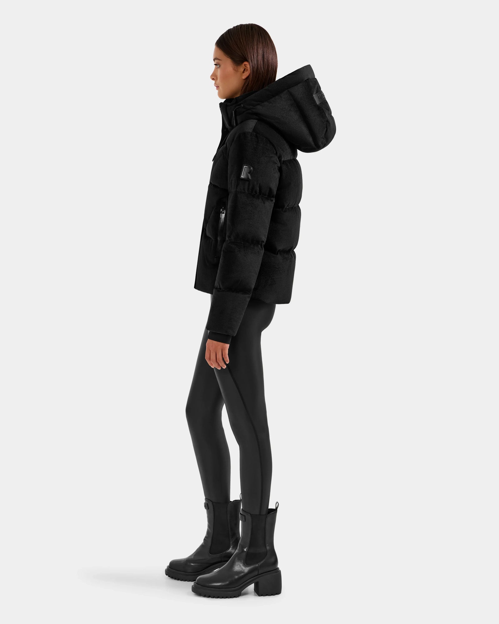 A woman stands in profile against a light gray background, wearing the Rudsak STEFANIE V - L125584 Black down puffer jacket, paired with black leggings and chunky-heeled black ankle boots.