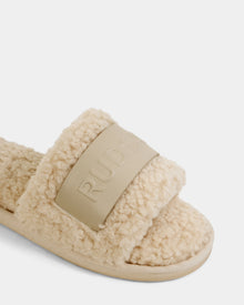 Close-up of the Rudsak TEDDY - L225517 Vanilla slide sandal featuring a beige, fleece-like design with a vegan-leather strap and an embossed RUDSAK panel—ideal for cozy loungewear looks.