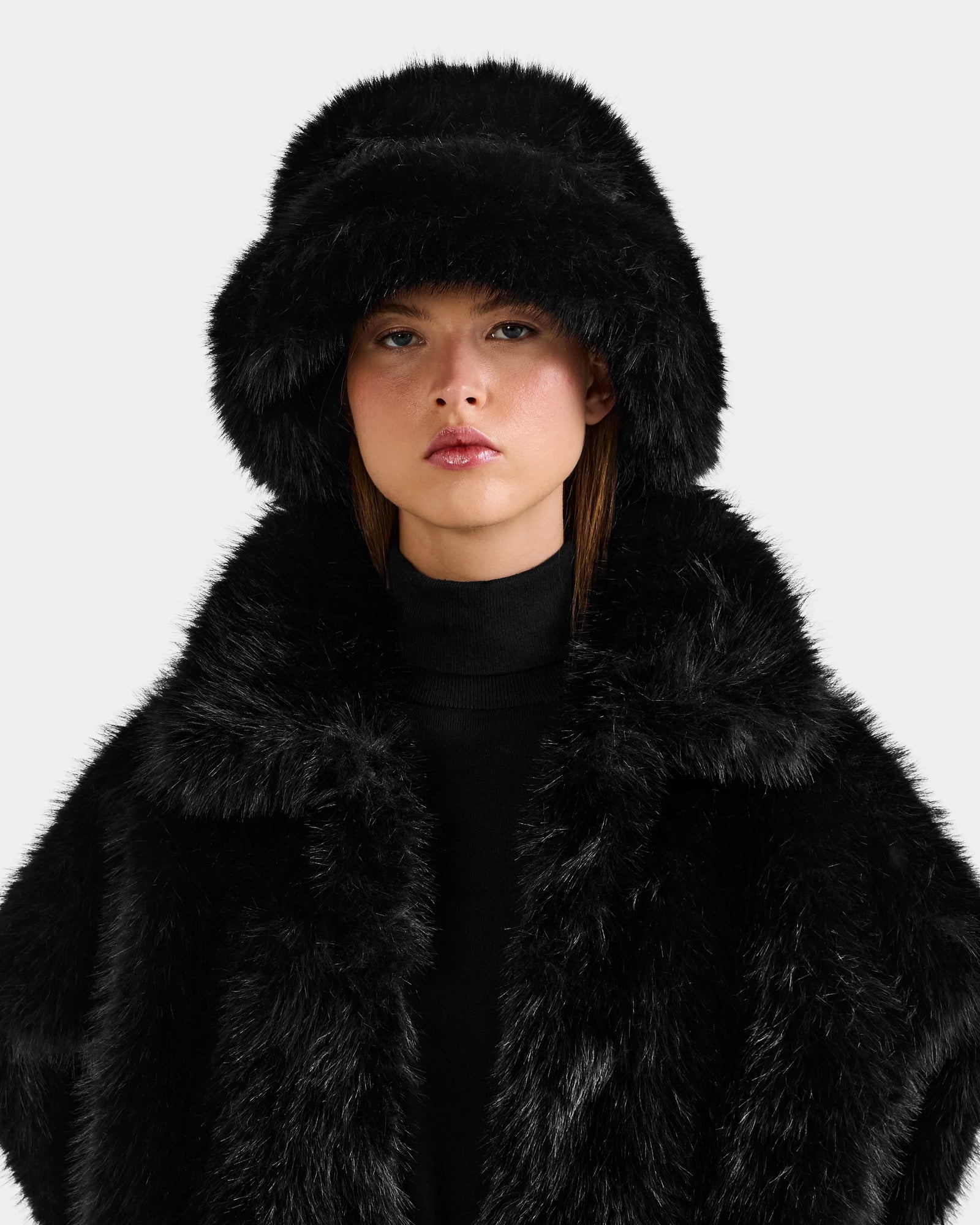 A person wears the Rudsak YURI - L425538 Black faux-fur bucket hat and matching coat with vegan-leather trim over a black turtleneck, facing forward with a neutral expression—an elegant winter accessory look for women.