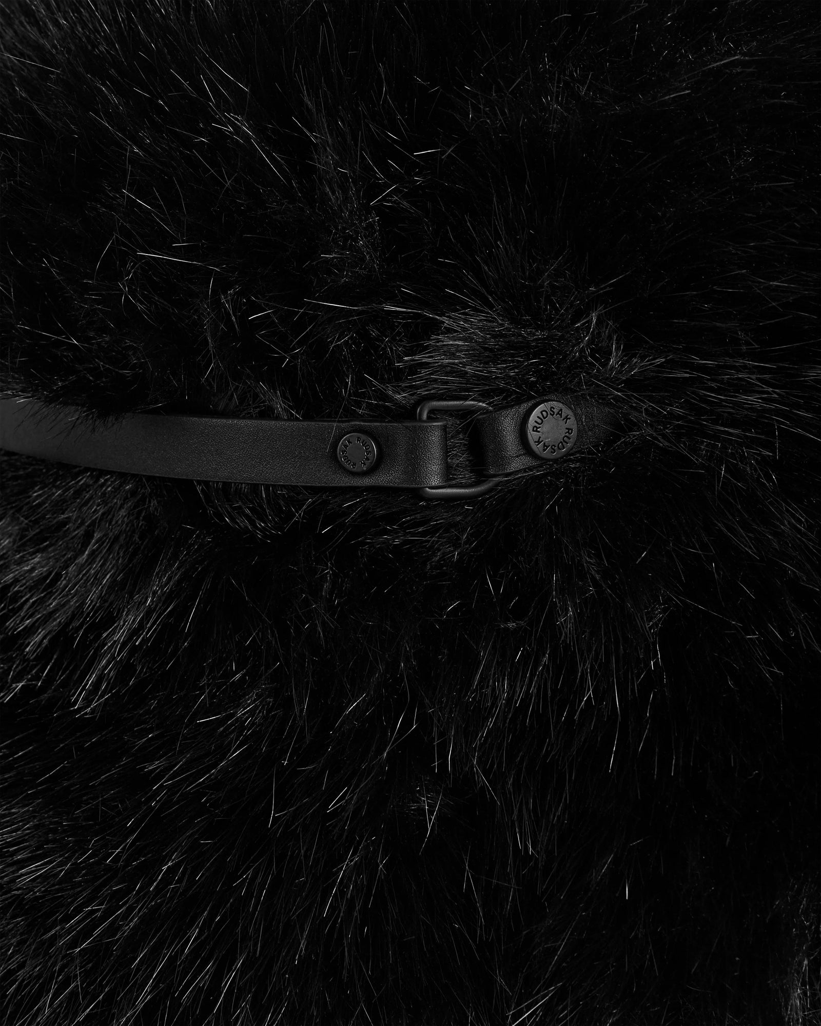 Close-up of Rudsak’s YURI - L425538 Black: black faux fur fabric with vegan-leather trim, a black leather strap, and metal rivets (one engraved), creating a textured, luxurious winter accessory.