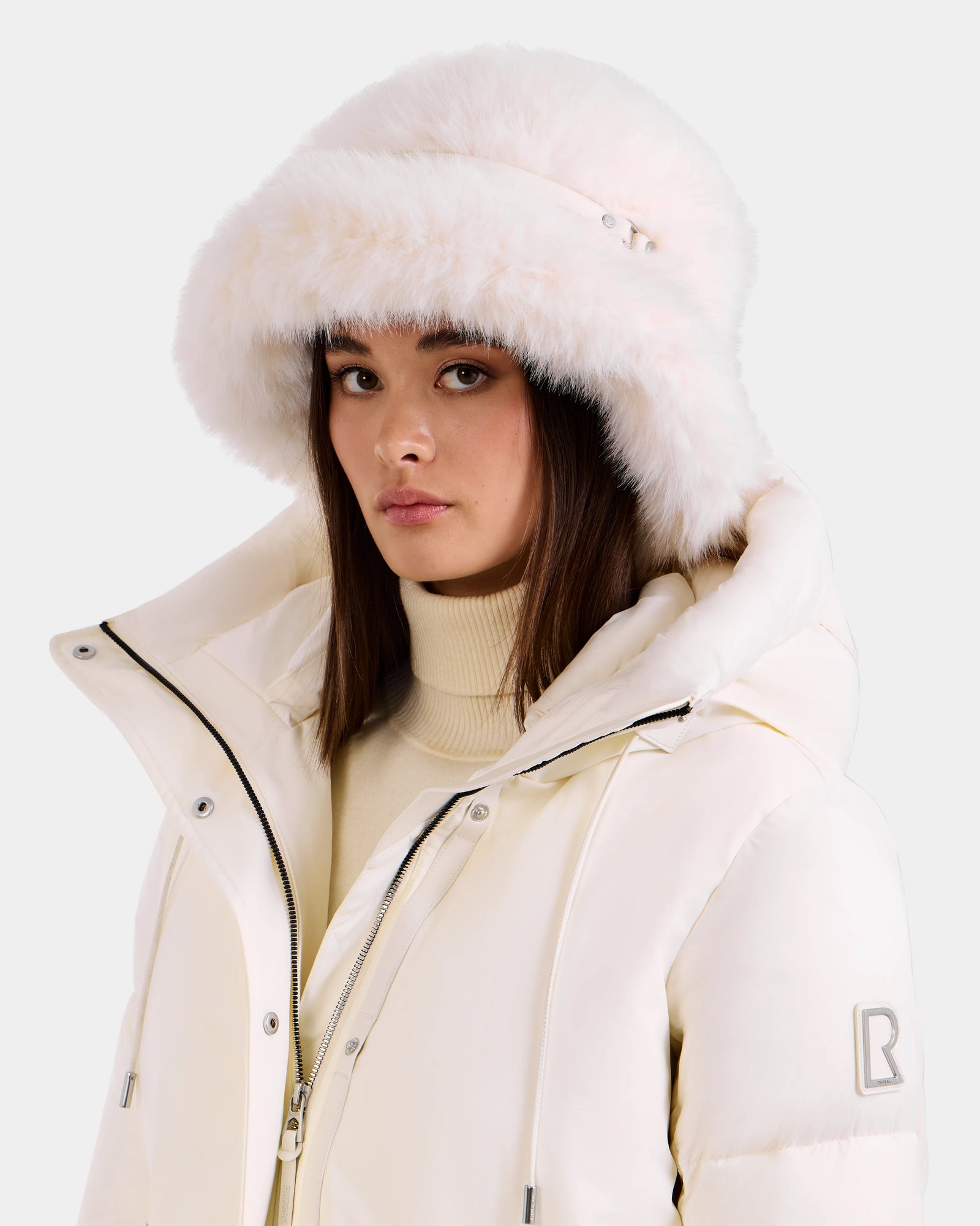A woman wears the Rudsak YURI - L425538 Iceland white puffer jacket with vegan-leather trim and a faux-fur bucket hat, posing against a light background—a chic winter look.