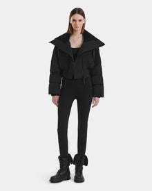 A woman faces forward against a white background, wearing the Rudsak YVETTE - L125557 Black cropped down jacket with an oversized collar, paired with black leggings and black combat boots. Her straight hair is down and her expression is neutral.