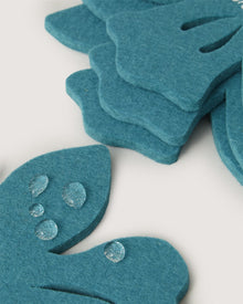 A close-up of Graf Lantz fabric shows teal, petal-like shapes crafted from premium German Merino wool, layered for texture. Clear droplets create a subtle gleam on the surface against a soft white background.