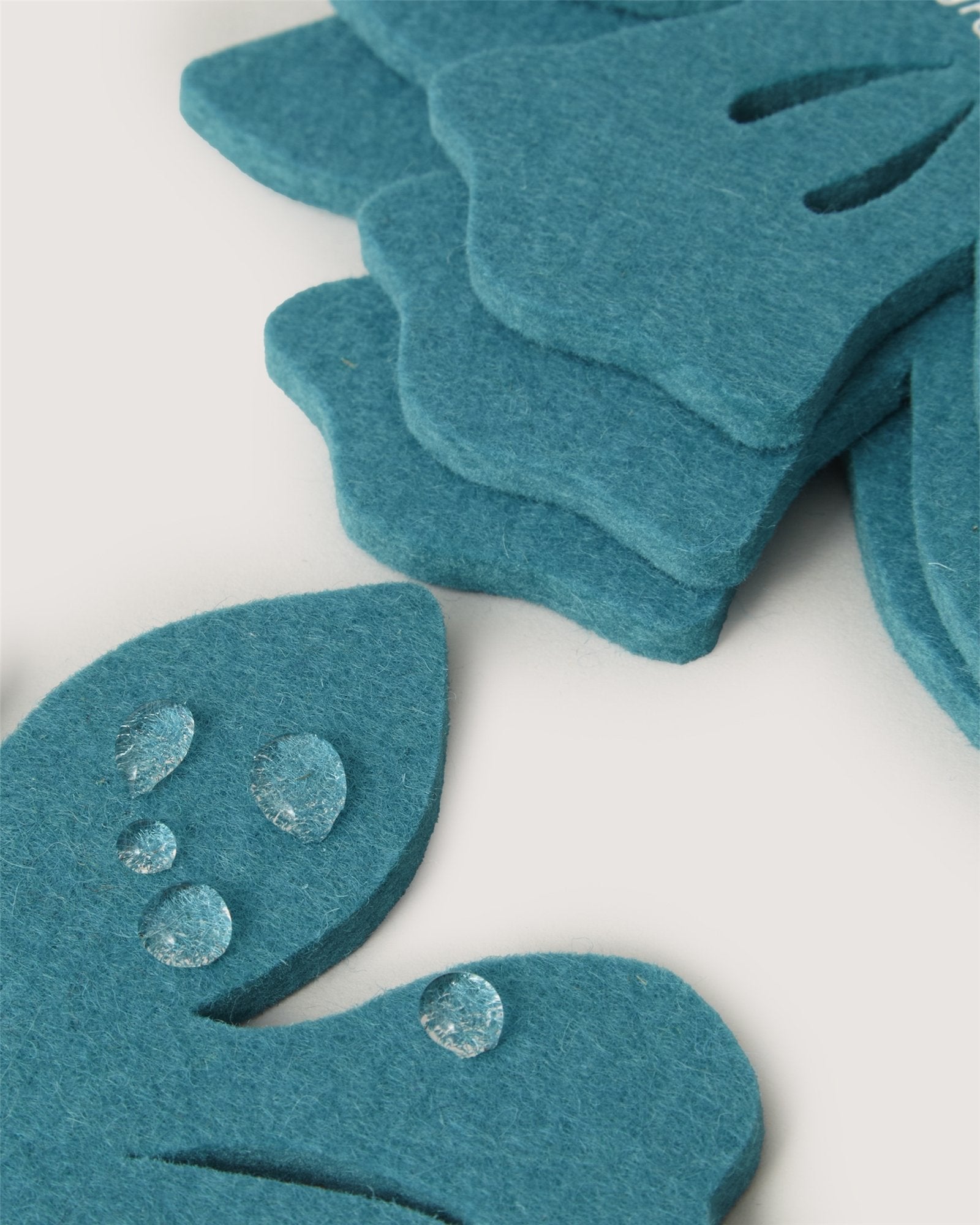 A close-up of Graf Lantz fabric shows teal, petal-like shapes crafted from premium German Merino wool, layered for texture. Clear droplets create a subtle gleam on the surface against a soft white background.