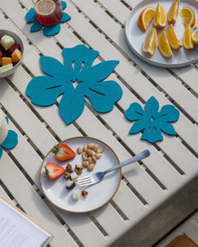 A white slatted table, decorated with blue floral place mats and Graf Lantz Merino wool coasters, showcases plates of mixed fruits, orange slices, nuts, and cheese. A glass of red drink with a lemon slice sits next to an open magazine.