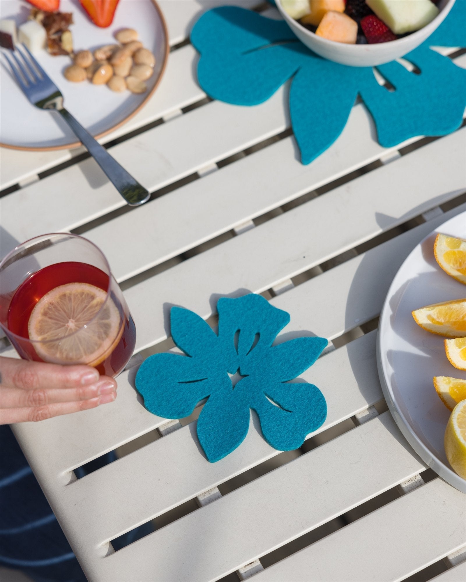 A hand holds a glass with a lemon slice on a table featuring teal floral placemats made from sustainable fabric. Plates with lemons, fruit, and snacks invite indulgence nearby, while Graf Lantzs Merino wool coasters add premium German charm.