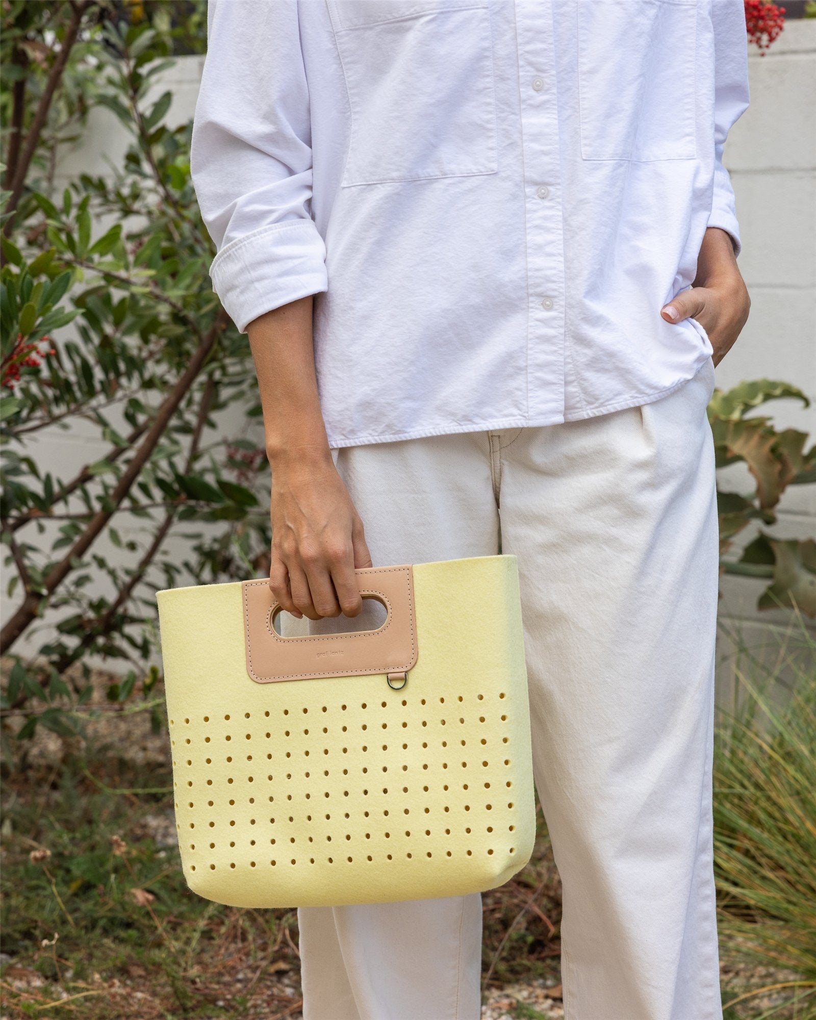 A person in a white outfit holds the Graf Lantz Asobi Bag, featuring perforated holes and a beige handle. Against lush greenery and a white wall, the light yellow tote beautifully complements their serene surroundings.