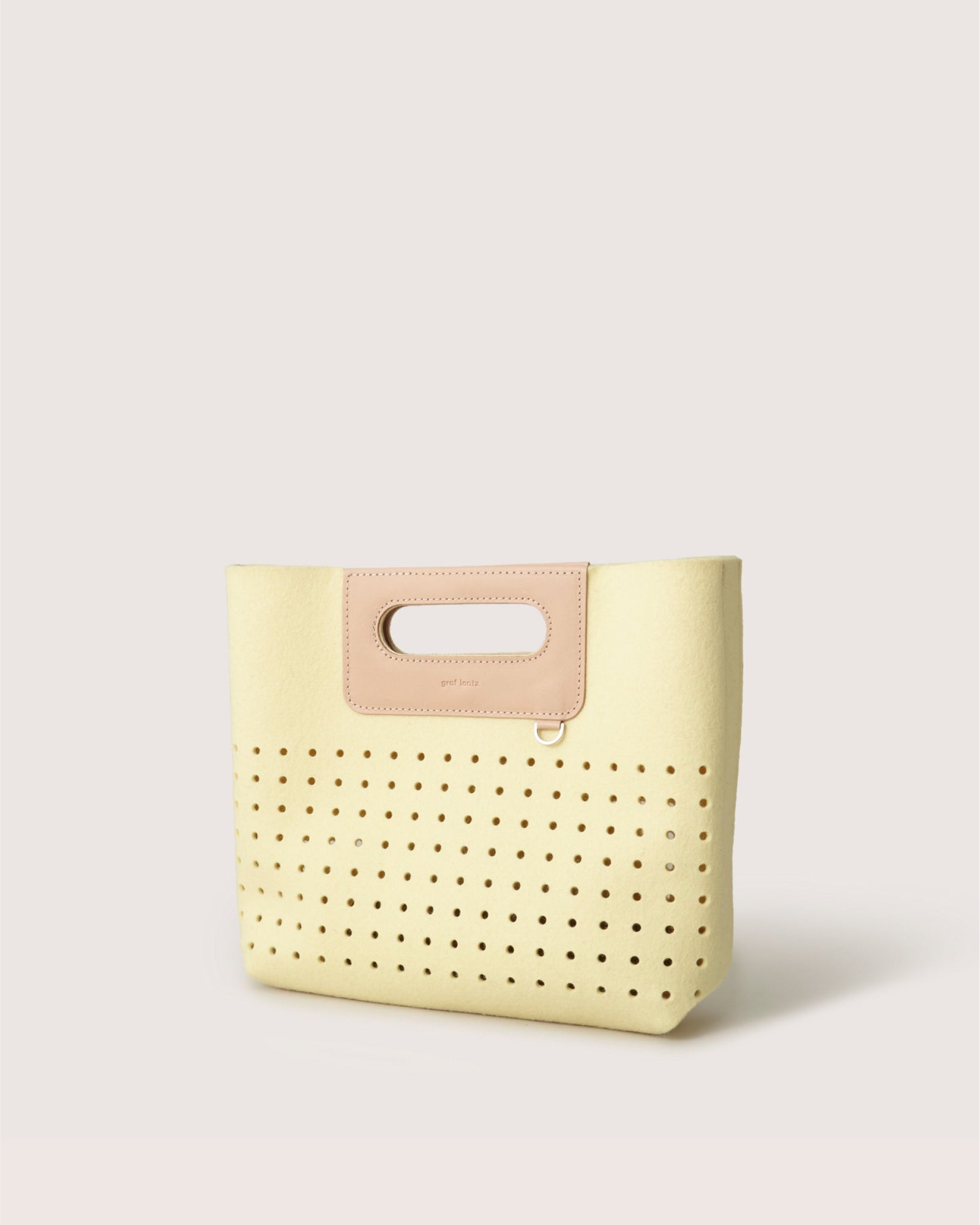 The Graf Lantz Asobi Bag, a minimalist handbag made from Merino wool, showcases a cream design with perforated holes and a tan leather handle cutout, elegantly set against a plain white background.