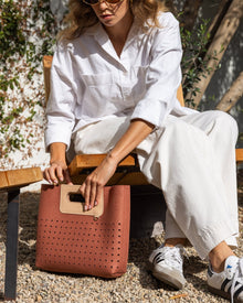 A woman sits on a wooden bench wearing a minimalist white outfit and sunglasses. She holds a Graf Lantz Asobi Bag, notable for its perforated design and rectangular handle. Small pebbles cover the ground as lush green foliage frames the serene scene.