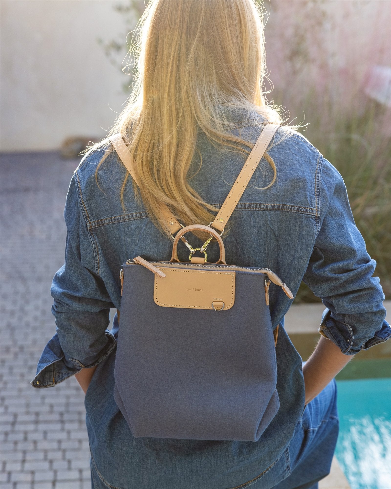 By the pool, a person with long blonde hair wears a blue denim jacket and displays a Graf Lantz multifunctional bag with beige straps. The scene includes outdoor tiles and greenery, reflecting their dedication to sustainable fashion.