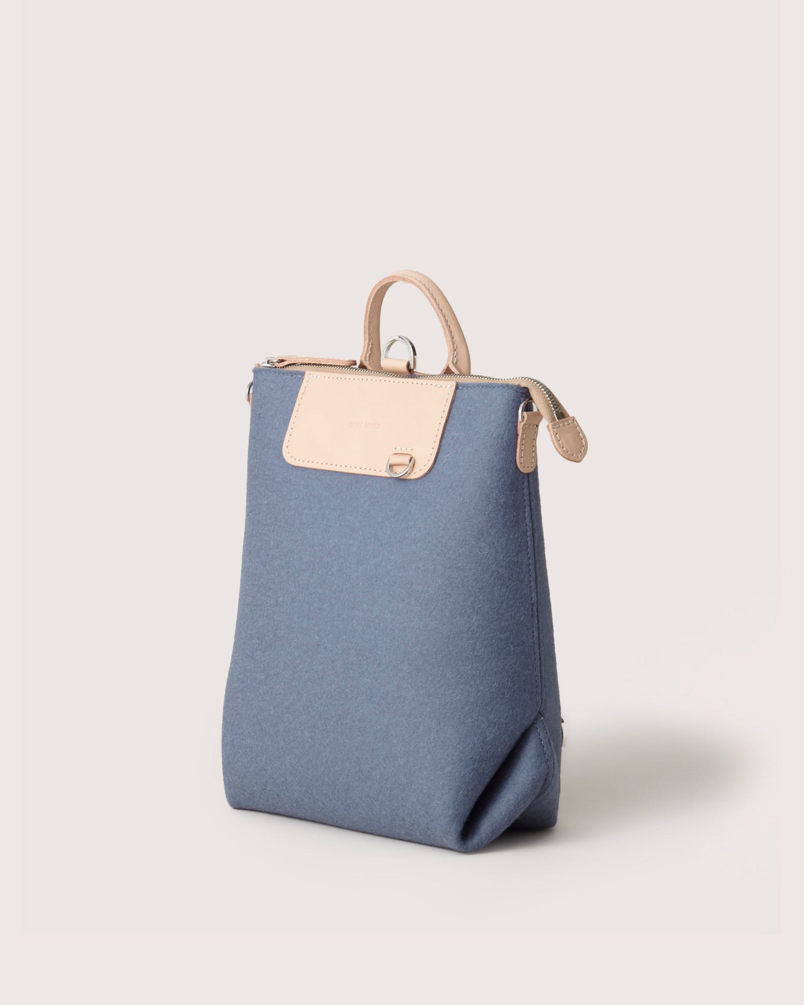 The Graf Lantz backpack features a stylish blue canvas with flap-over closure and tan leather accents on the top handle and flap. This minimalist design includes a subtle tan emblem on the flap and is captured against a plain background.