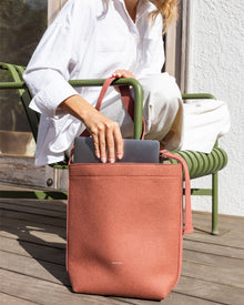 A person in a white outfit slips a laptop into a large, salmon-colored Graf Lantz tote bag with any tote printed on it. This Merino Wool Tote offers sustainable functionality as they relax on an outdoor green chair, with part of the door and wall visible behind.