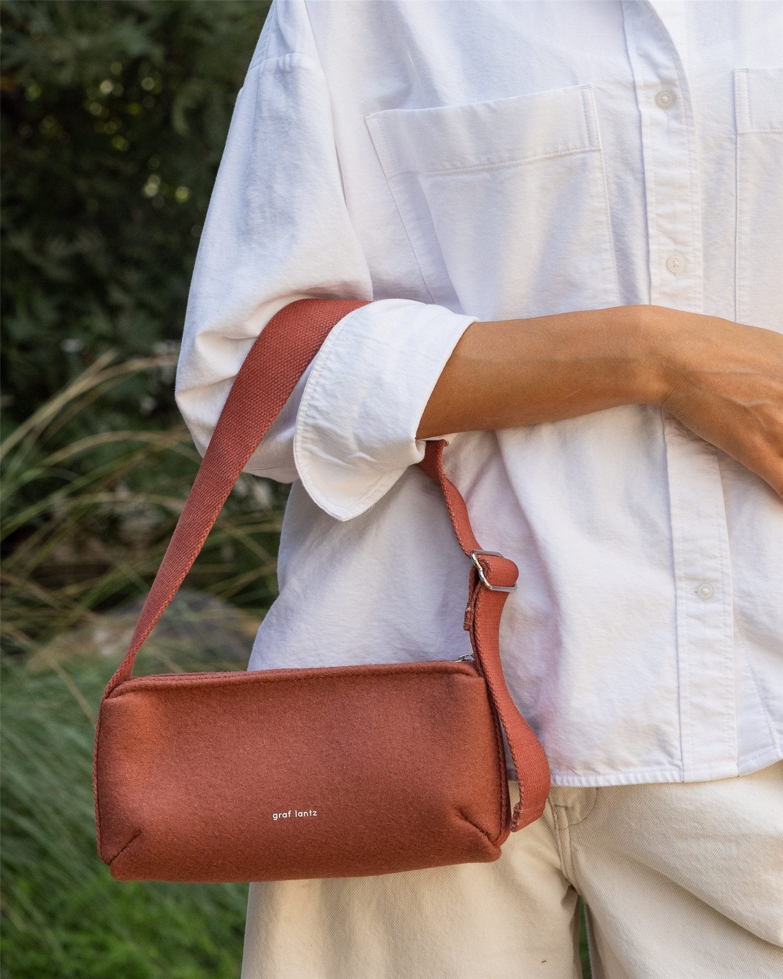 A person in white and light pants holds a rust-colored Graf Lantz Campus Shoulder Bag, featuring impressive fonts. The scene is set against a lush green backdrop.