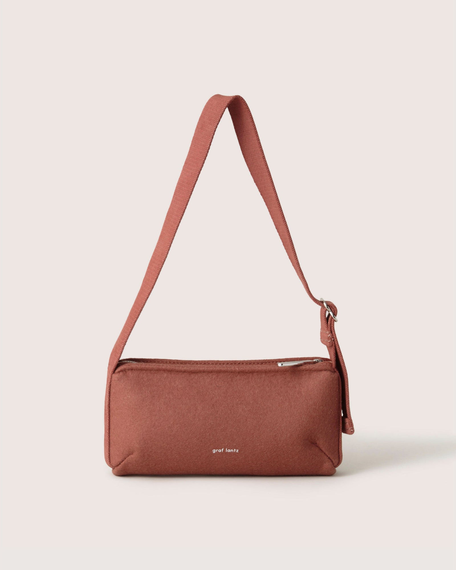 The Graf Lantz Campus Shoulder Bag, a rust-colored rectangular accessory made of soft Merino Wool, holds everyday essentials. It features a zippered top, single strap, and subtle white branding.
