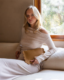 A woman with long blonde hair sits on a cream sofa in an off-shoulder beige sweater dress, holding a versatile Graf Lantz clutch with deco edges. She gazes softly at the camera as sunlight filters through a window, revealing lush greenery outside.
