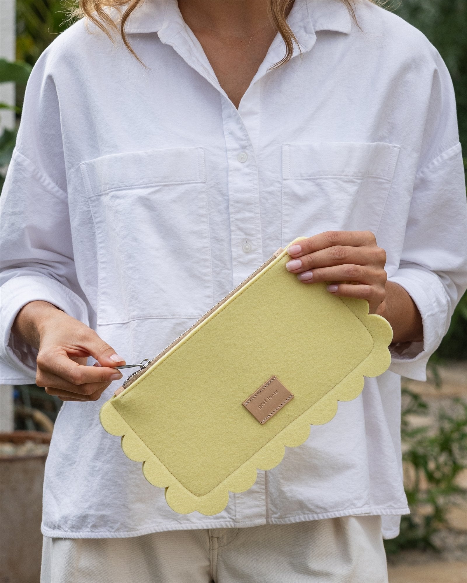 A person in a white shirt holds the spacious yellow Deco Edge Pouch by Graf Lantz, featuring scalloped edges and a small tan label, against a backdrop of blurred greenery.