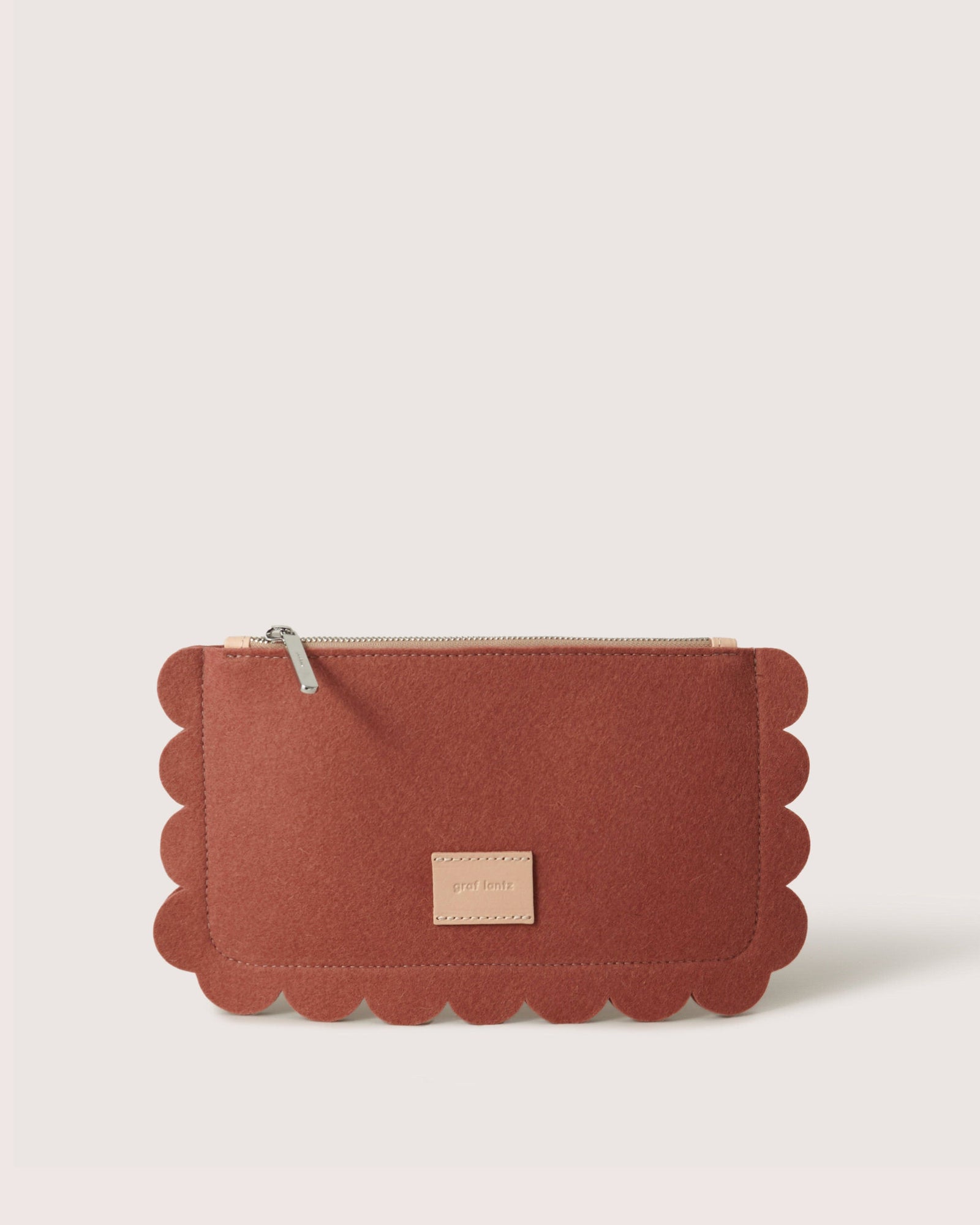 The Graf Lantz Deco Edge Pouch, crafted from sustainable Merino wool, is a rectangular rust-colored felt pouch. It features a scalloped edge, visible zipper, and a small beige label on its plain light-colored front.
