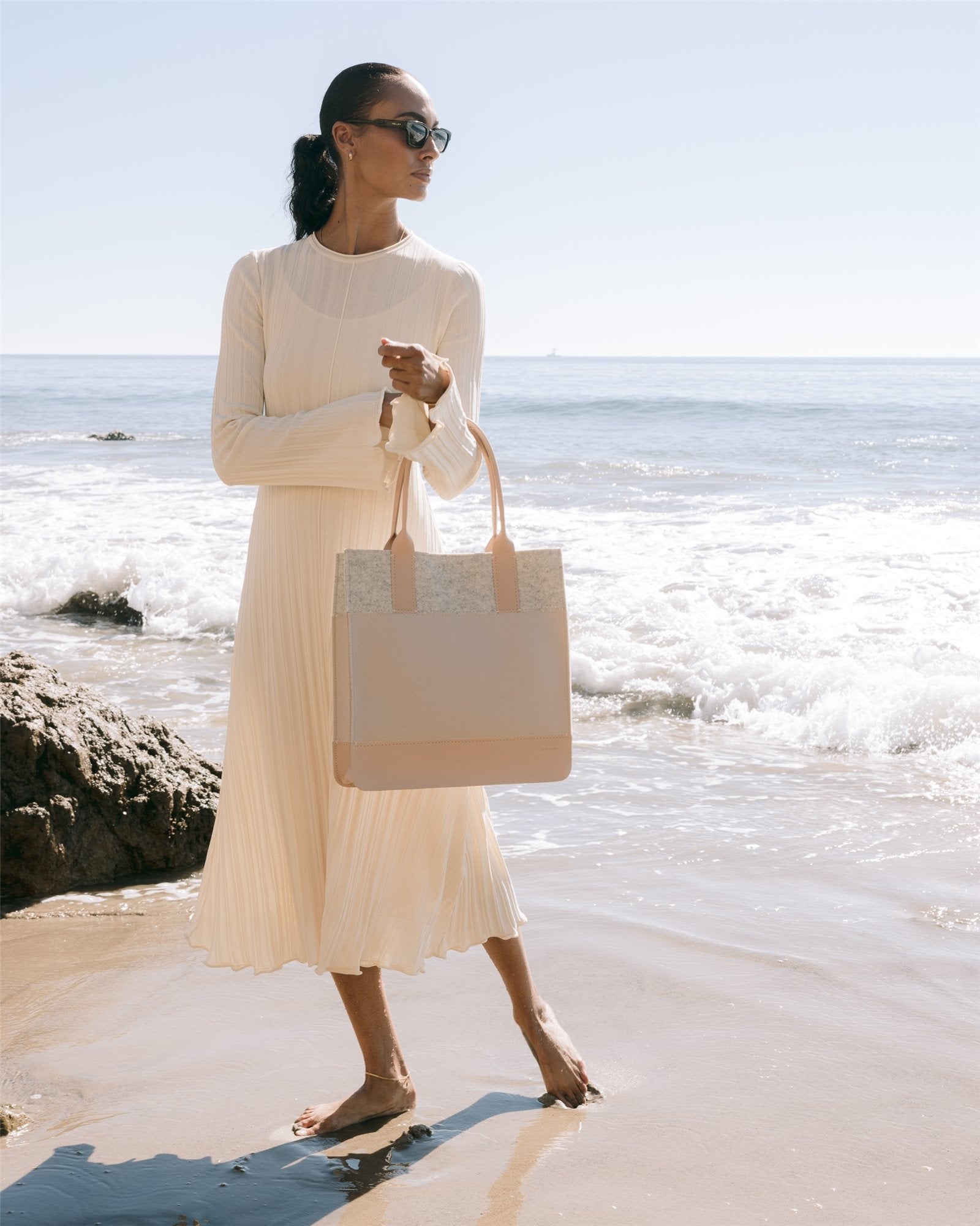 Barefoot on a sandy beach under a clear blue sky, a woman in a long-sleeved cream dress holds her Jaunt Midi Tote by Graf Lantz, wearing sunglasses and gazing into the distance as waves gently lap at the shore.