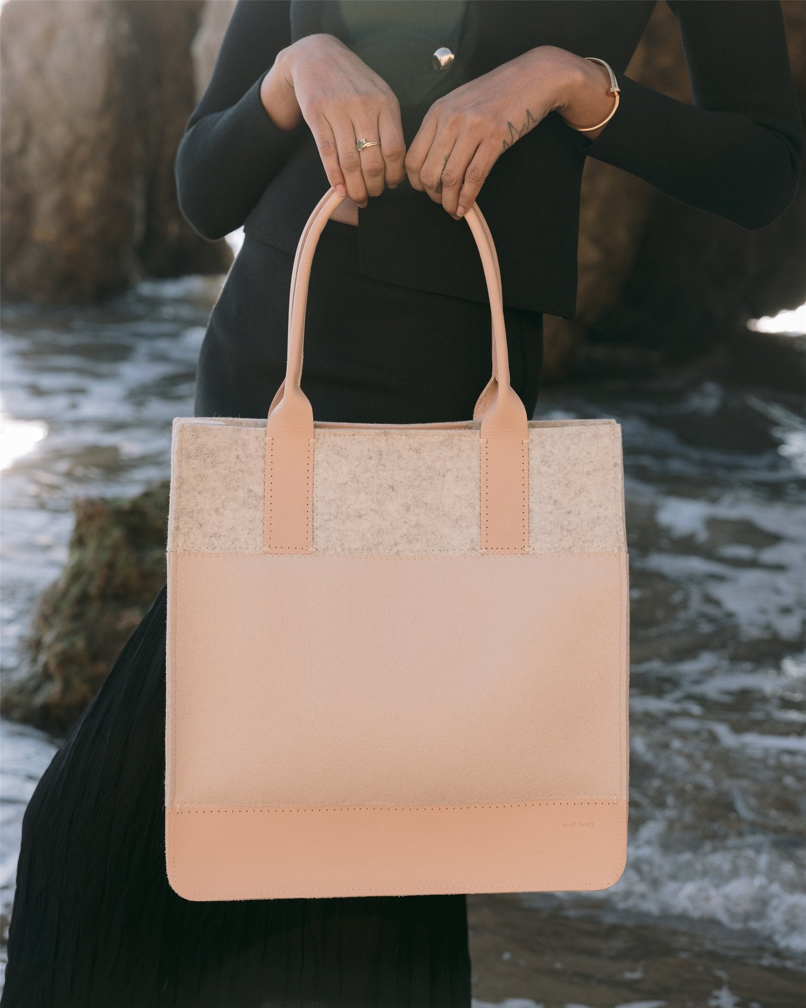 A person dressed in black stands by a rocky shore, holding the stylish Graf Lantz Jaunt Midi Tote with light fabric accents, enjoying the ocean waves in the background.