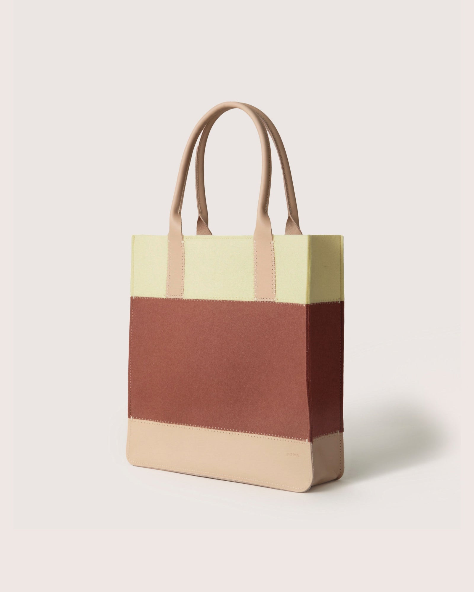 The Graf Lantz Jaunt Midi Tote is a mid-size bag with dual handles, featuring three trendy colors: muted yellow at the top, brown in the middle, and beige at the base. It stands stylishly upright against a simple light background.