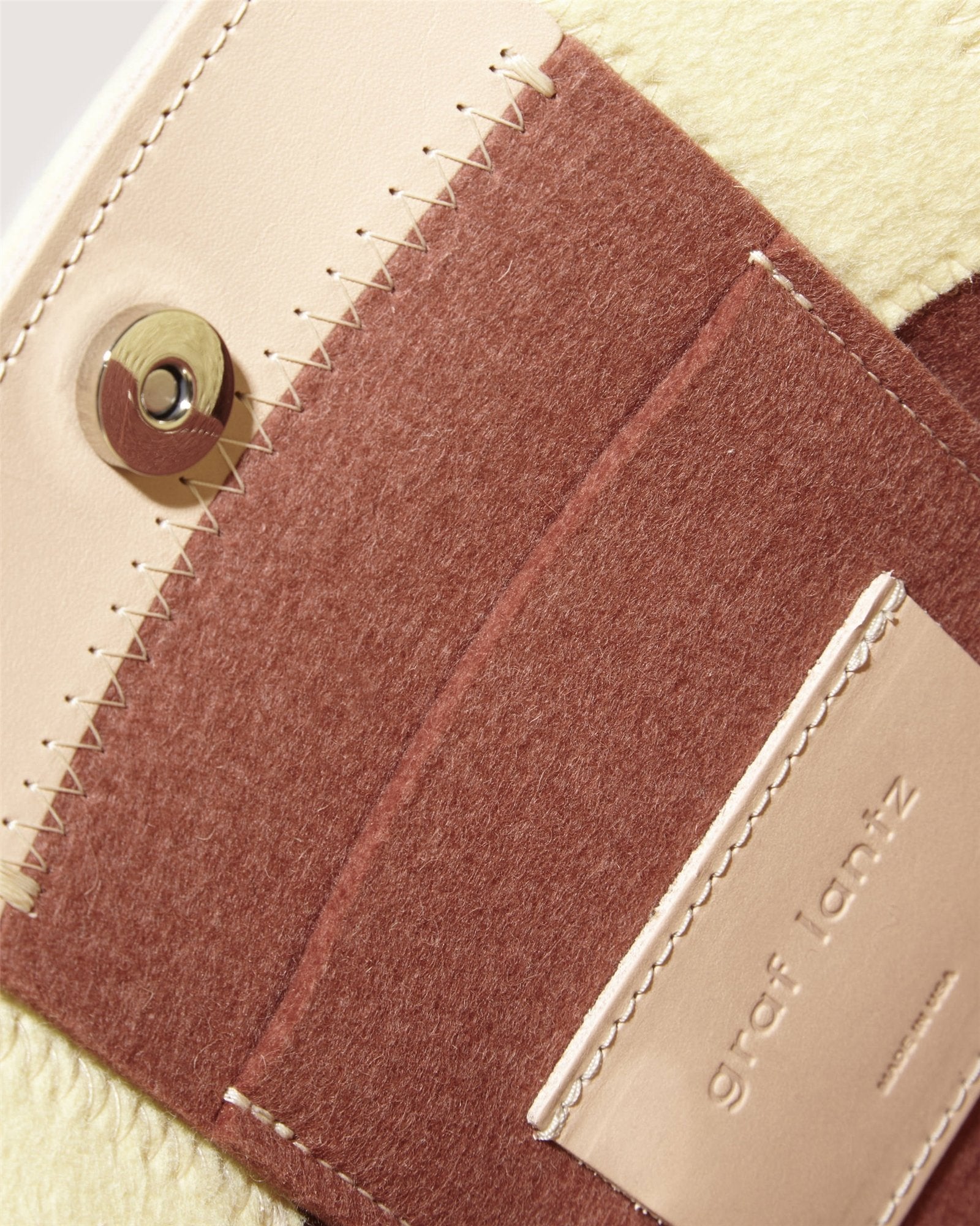 Close-up of a textured Merino wool fabric item with stitched brown and beige pockets. A round metal button is near the top, with a Graf Lantz embossed leather patch on one pocket, resembling details found on the Jaunt Midi Tote.
